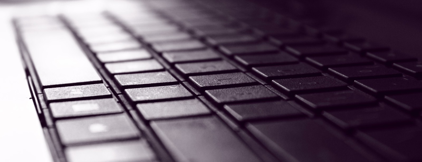 Rows of dark laptop keys recede into soft shadow, their edges catching pale light from one side.
