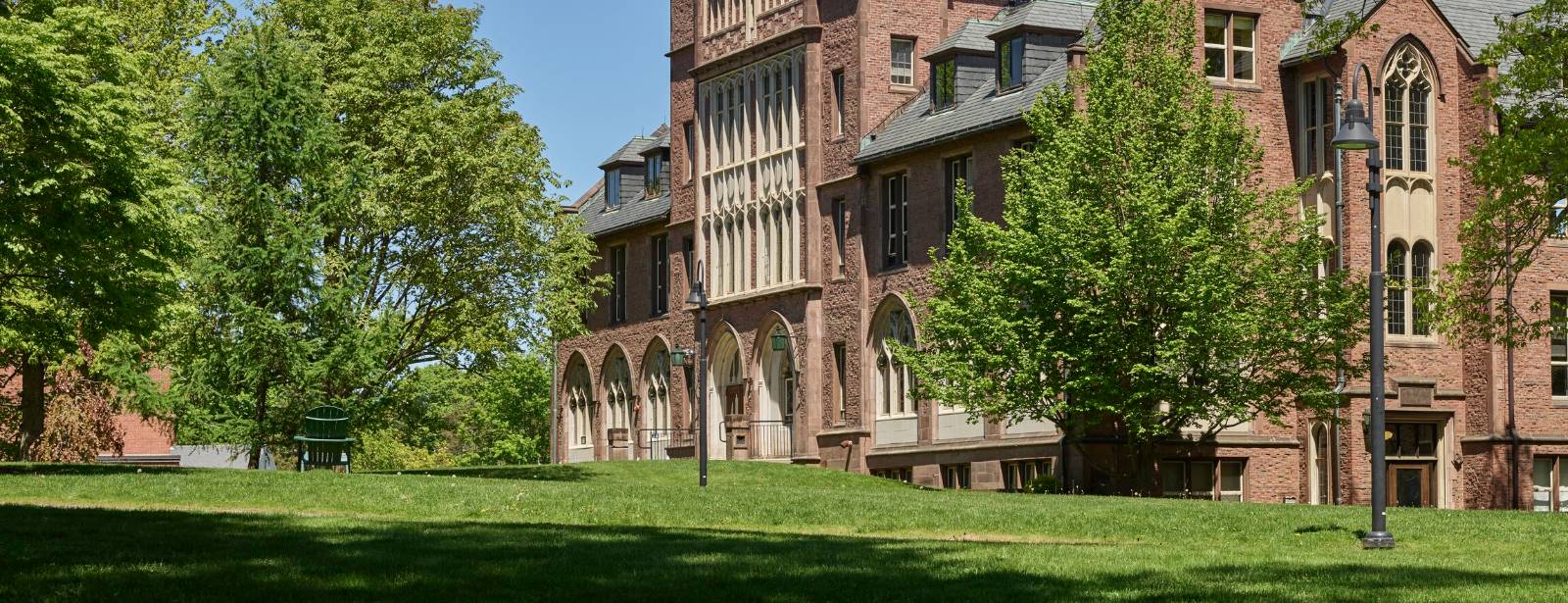 A brick and stone hall with arched doorways and tall Gothic windows stands behind trees across a sunlit lawn.