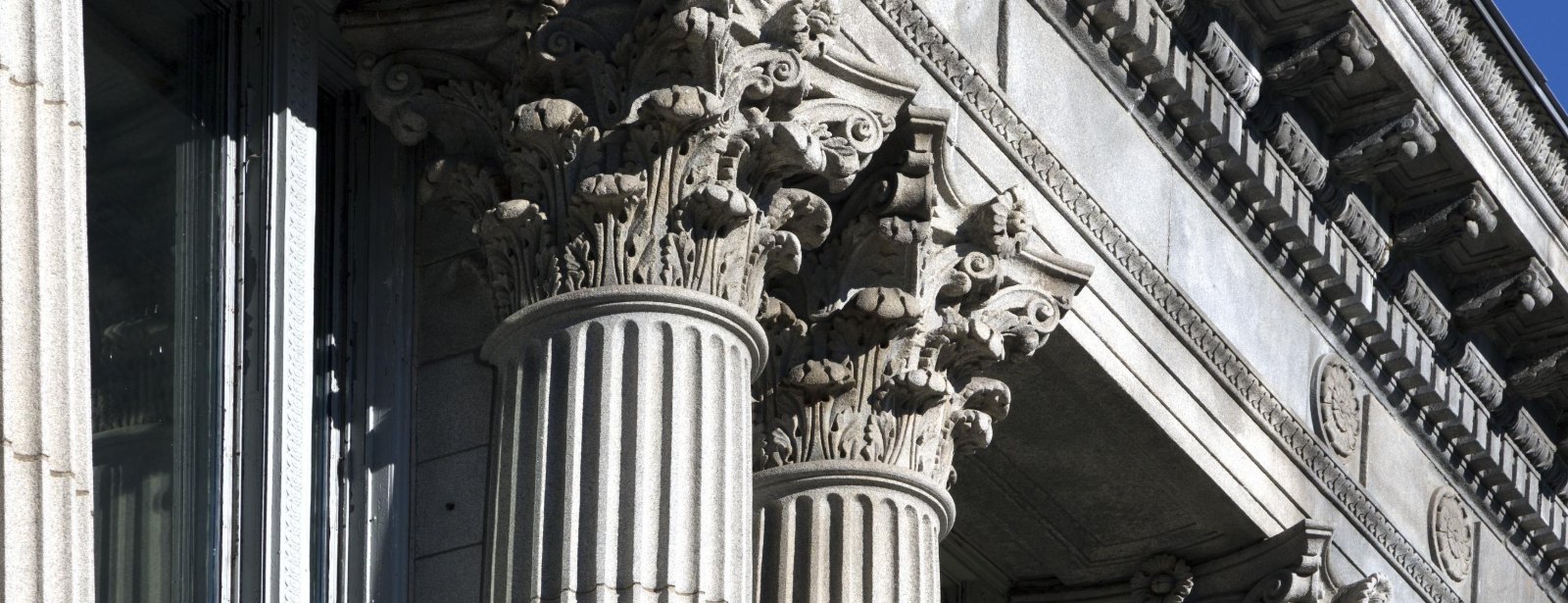 Fluted stone columns topped with carved Corinthian capitals stand in bright sun beneath a deeply shadowed cornice.