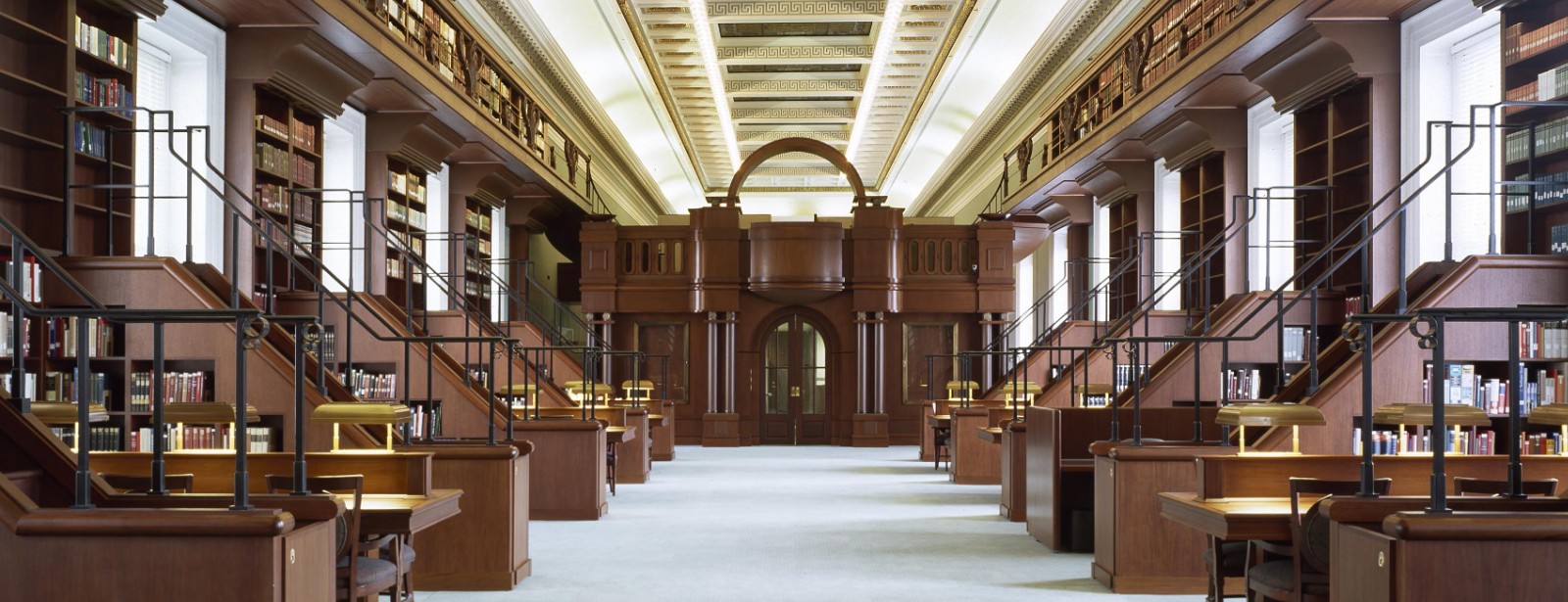 Wooden reading desks and book-lined galleries run the length of an empty library hall toward an arched wooden doorway.