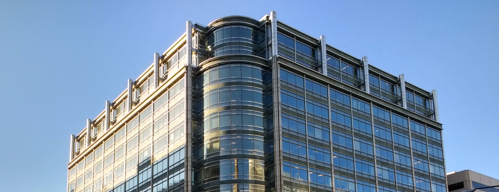 Sunlight catches the glass and steel corner of an office building, with a curved glass bay rising against a clear sky.
