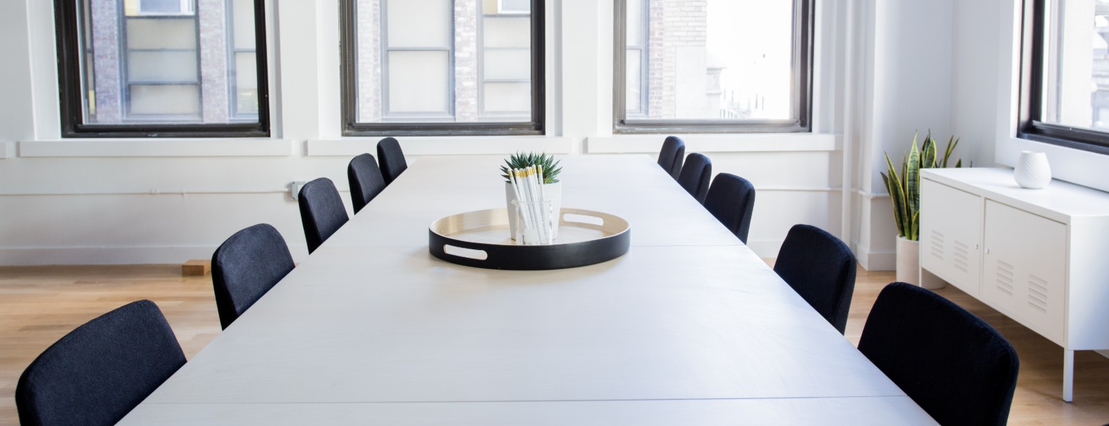 A long white table with black chairs runs toward tall windows in a bright, empty meeting room.