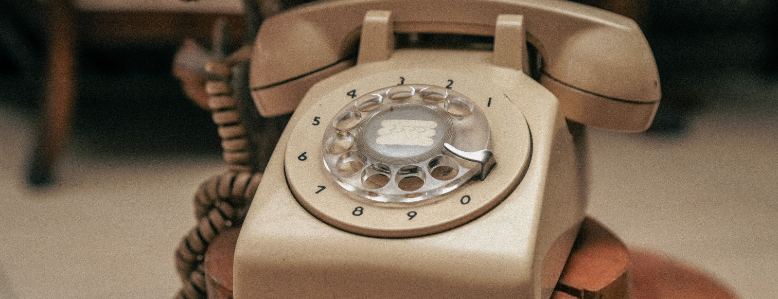 A beige rotary telephone with a coiled cord rests on a wooden stand, its handset in the cradle.