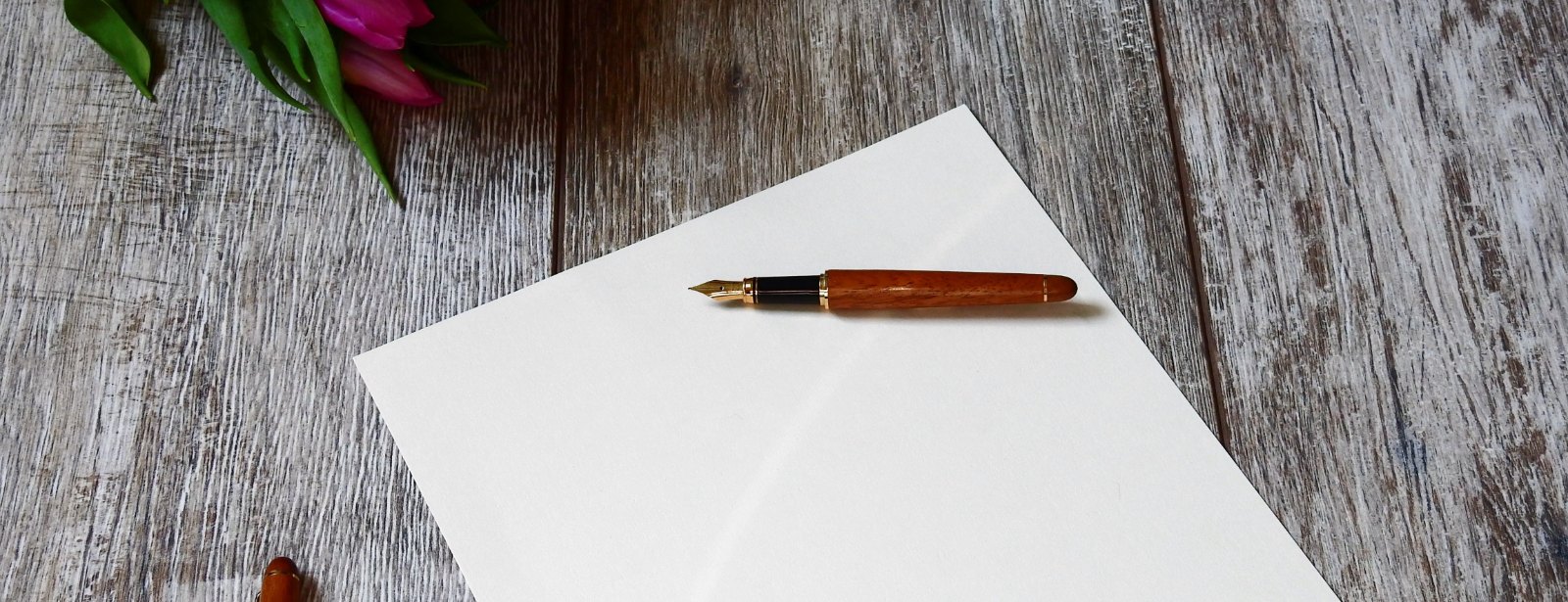 A fountain pen with a wooden barrel lies on a blank white sheet of paper on a weathered wooden table.